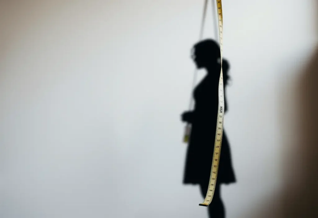 Un silhouette floue se tient près d’un mur, un ruban souple pend à côté, illustrant la prise de mesure du tour de poitrine.