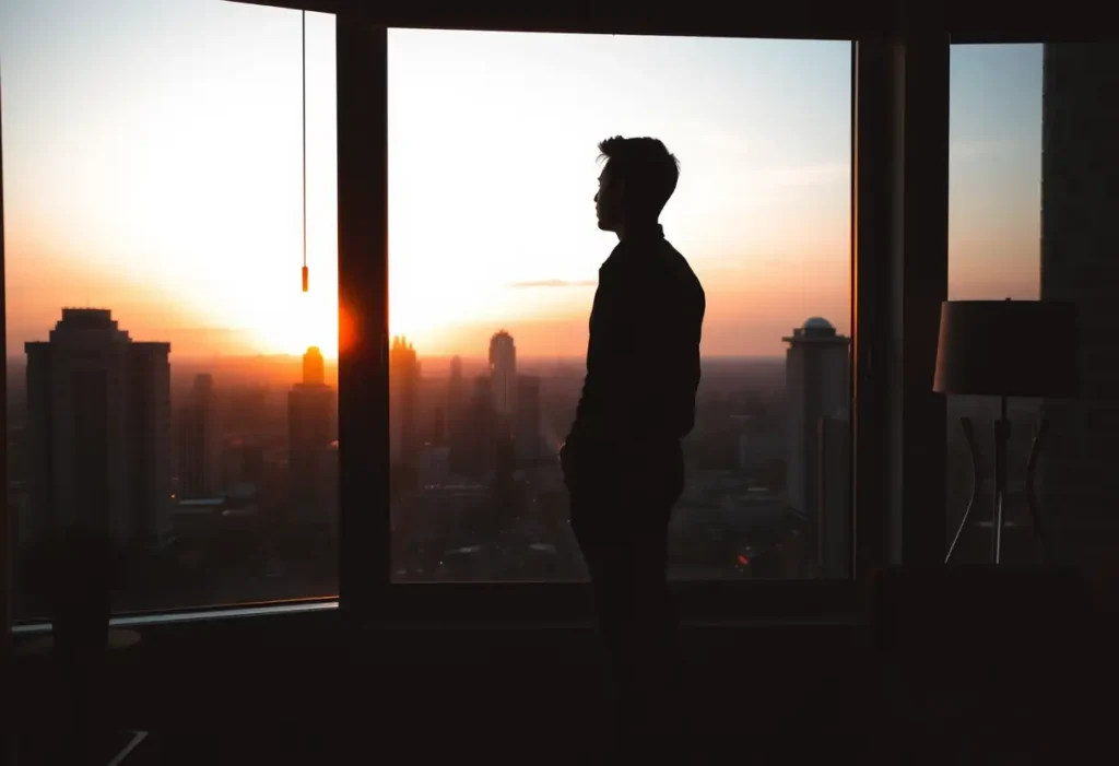 Un silhouettage de personne près d'une fenêtre dans un appartement moderne, regardant une skyline de ville au coucher du soleil