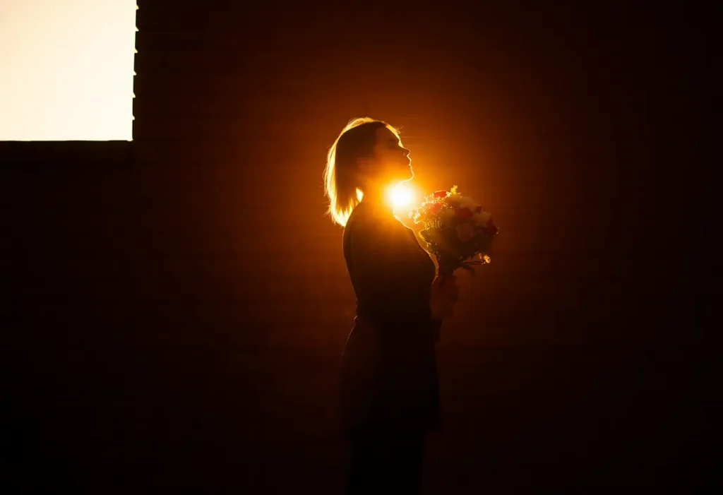 Un profil silhouetté contre un mur de brique, tenant une gerbe de fleurs sous la lumière du soleil couchant, illustrant l'originalité des gestes d'affection.
