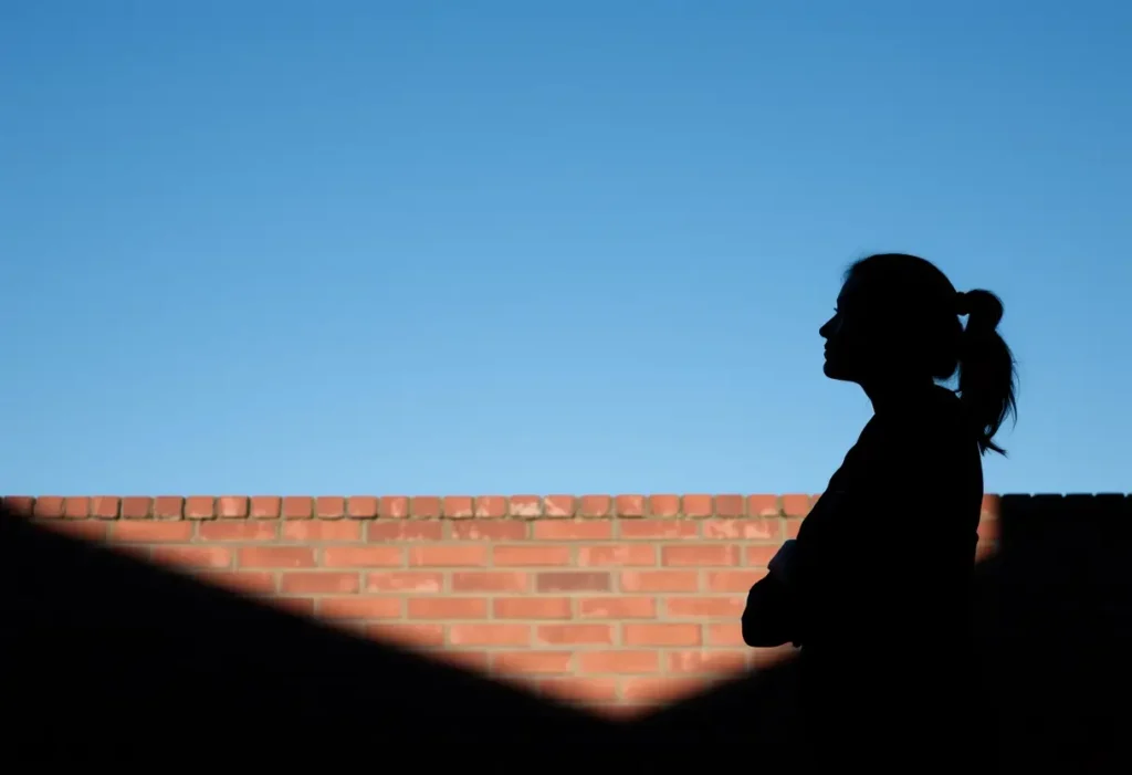 Un profil flou d'une femme aux bras croisés se reflète contre un mur en brique sous un ciel bleu clair, elle réfléchit à sa croissance personnelle et son émancipation